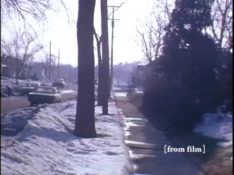 The still captures a wintertime suburban street with snow lining the sidewalk and two men casually conversing by a classic car parked nearby. The camera angle is positioned at a slight distance, emphasizing the serene neighborhood atmosphere framed by bare trees and distant homes.