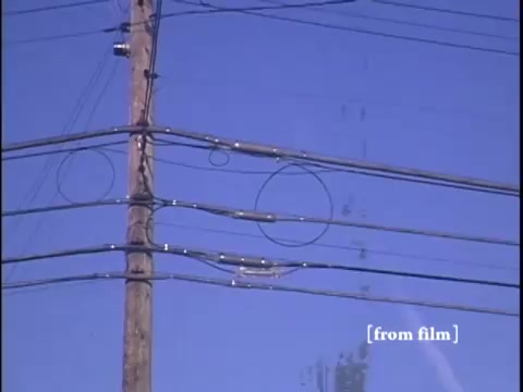 The still captures a winter suburban street scene with two men near a car, framed by a bright blue sky and the intricate details of utility poles and wires. In the foreground, a yellow fire hydrant adds a splash of color against the muted tones of the setting.