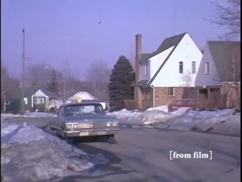 The film still captures a wintry suburban street scene featuring two men near a parked car, with a yellow fire hydrant visible and snow piled along the road. The camera is positioned at a low angle, emphasizing the residential homes and the suburban environment.
