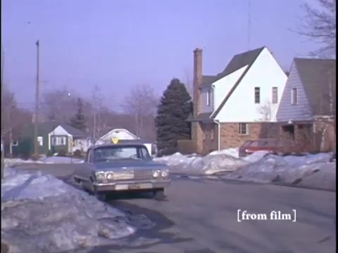 The still captures a winter suburban street scene featuring a parked car and two men, with patches of snow lining the road. The camera angle is positioned at street level, showcasing homes in the background, including a prominent yellow fire hydrant.