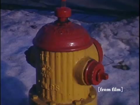 The film still features a close-up of a vibrant yellow fire hydrant set against a snowy suburban street in winter. In the background, hints of a car and the facade of a nearby home suggest a quiet neighborhood atmosphere.