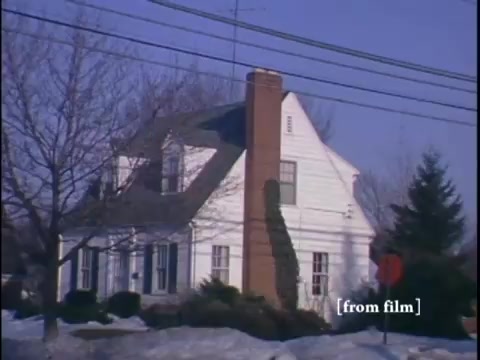 The still captures a wintertime suburban scene, showcasing a white house with a prominent chimney framed by leafless trees and a stop sign. In the background, a glimpse of a car and two figures can be seen on the street, under a clear blue sky.