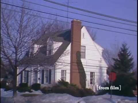 The still captures a quiet winter scene on a suburban street, featuring a white house framed by leafless trees and a yellow fire hydrant nearby. The camera angle, positioned from across the street, highlights the charming residential architecture and the serene atmosphere of the neighborhood.