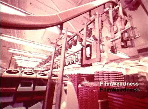 The still from the 16mm film 'Bell Performance' showcases a manufacturing environment from the 1970s. It features an assembly line where telephones are being produced, with various components such as phone casings and parts suspended from overhead tracks. The imagery reflects a nostalgic glimpse into the industrial processes of the Bell System, characterized by a vibrant color palette and a focus on efficiency in telecommunications manufacturing.
