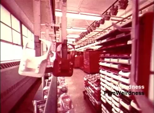 The still from the 1970s Bell System film 'Bell Performance' depicts an industrial setting focused on telephone manufacturing. The scene shows an assembly line with partially assembled telephone components suspended from conveyor belts, surrounded by organized racks of materials. The environment appears to emphasize efficiency and automation, typical of manufacturing practices of that era. The colors and aesthetic evoke a sense of nostalgia, highlighting the technological advances of the time in telecommunication production.