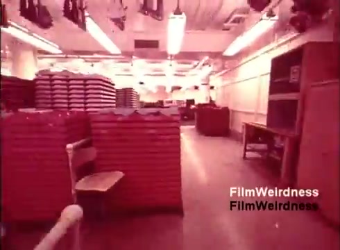 The still from 'Bell Performance' depicts a large, industrial workspace, likely from the 1970s, characterized by rows of stacked materials, possibly telephone components. A lone chair is positioned in the foreground, indicating the working environment. The overall atmosphere is nostalgic, with a warm, reddish tint that evokes a sense of historical significance associated with the Bell System's telecommunications legacy. The absence of titles suggests a focus on the manufacturing process rather than storytelling.