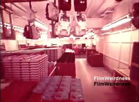 The still from 'Bell System Film from the 1970s' showcases a manufacturing environment, likely depicting the assembly of telephones. It features overhead machinery, possibly robotic arms, moving in a factory setting. There are stacks of materials and components organized on shelves, indicative of an efficient production line. The color tone appears warm, contributing to a nostalgic feel. This scene captures a moment in the technological evolution of communications, highlighting the Bell System's role in telephone manufacturing during that era.