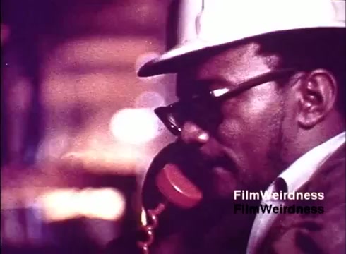 The still from the 'Bell System Film from the 1970s' features a man wearing a hard hat and glasses, engaged in a phone conversation. The setting appears industrial, likely within a manufacturing facility focused on telecommunications. This captures a moment of hands-on work and emphasizes the human element in the production of telephones, reflecting the era's connection to technological advancement and the Bell System's legacy. The color tone adds a nostalgic feel to the scene.