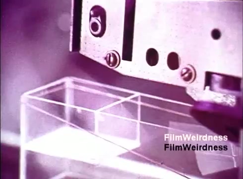 The still from 'Bell System Film from the 1970s' shows a close-up of a mechanical process involved in the manufacturing of telephones. The image captures a machine apparatus, possibly a robotic arm, hovering above a transparent, rectangular plastic component. The overall aesthetic has a vintage feel, with a muted color palette that includes shades of purple, suggesting it may have been created using older filming techniques. This conveys a sense of nostalgia, reflecting on the technological innovations of the time in the telecommunications industry.