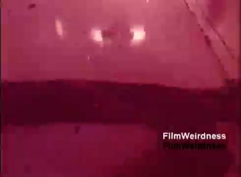 The still from the film 'Bell Performance' showcases a scene reminiscent of the 1970s, focusing on the manufacturing process of telephones. The colors appear muted, with a reddish tint dominating the frame, evoking a nostalgic, vintage aesthetic. The imagery captures the industrial atmosphere of a bygone era, highlighting machinery and possibly assembly line activity associated with the Bell System. Overall, it reflects a moment in time dedicated to the production of telecommunications equipment.