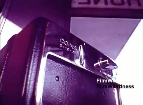 The still from the 16mm film 'Bell Performance' features a close-up of a classic payphone with a chrome finish, highlighting the coin slot labeled for payment. The background suggests a retro environment associated with telecommunications. The overall aesthetic reflects the design and technology of the Bell System era in the 1970s, evoking nostalgia for that period of innovation in phone technology.