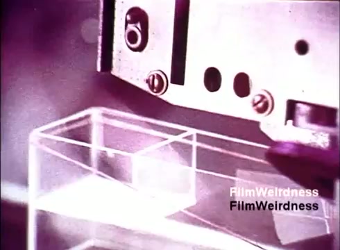 The still from 'Bell Performance' features a close-up of machinery involved in the manufacturing process of telephones. The focus is on a robotic or automated arm, highlighting the precision and technology of the era. The background suggests a factory or production environment, emphasizing the industrial nature of telephone assembly. The overall presentation reflects a nostalgic look at telecommunications innovation from the 1970s.