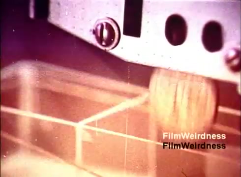 The 16mm still from the film 'Bell Performance' showcases a close-up of a manufacturing process, likely involving the assembly or testing of telephone components. The image captures mechanical apparatuses in motion, highlighting the intricate details of production. The warm tones suggest a vintage aesthetic, reflective of the 1970s era, evoking a sense of nostalgia associated with the Bell System and its telecommunications advancements.