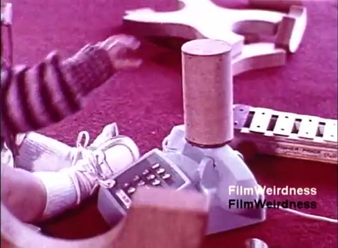 The still from 'Bell System Film from the 1970s' depicts a scene likely focused on a child's interaction with a toy telephone. The image features a small child wearing white shoes and a striped outfit, reaching toward a toy phone that resembles a classic model. The background seems to have various colorful objects, drawing attention to the playful environment. This snapshot embodies mid-20th-century nostalgia for telecommunications, highlighting the familial and educational aspects of learning through play.