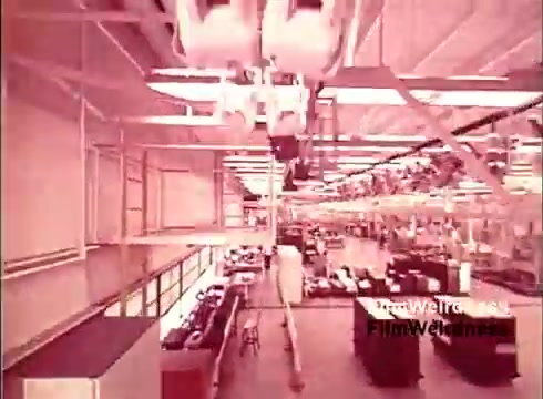 The still from 'Bell System Film from the 1970s' showcases a factory interior focused on the production of telephones. It features industrial machinery and assembly lines, highlighting the manufacturing process in a nostalgic red-tinted aesthetic. The environment appears bustling, indicative of a time when telecommunication technology was rapidly evolving.