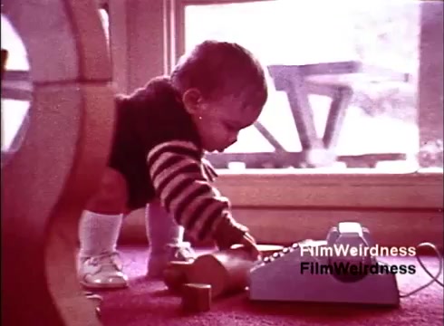 This still from the 1970s film 'Bell Performance' features a toddler in a cozy indoor setting, focused on a classic telephone. The child wears a striped sweater and white socks while kneeling on a vibrant carpet. The scene captures a moment of curiosity and exploration, evoking nostalgia for the era of telecommunication. The background suggests a warm, inviting home environment, contributing to the film's charm.