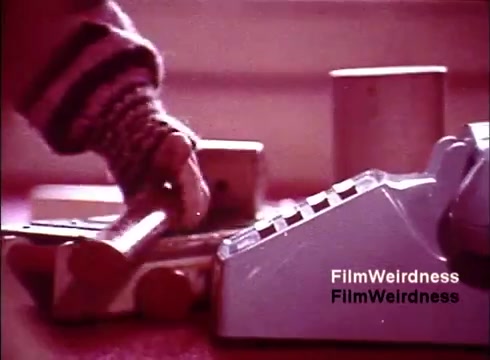 The still from the 1970s 'Bell System Film' titled 'Bell Performance' features a close-up shot of a hand interacting with a telephone. There are various objects, including a phone with a keypad, alongside what appear to be components or tools for assembling or repairing equipment. The color palette is warm, evoking a nostalgic atmosphere. This moment captures the era's focus on telecommunications and the innovation surrounding the manufacturing of phones.