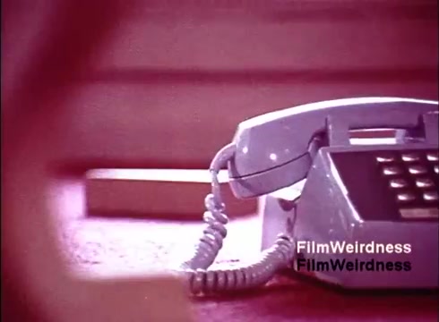 The still from the 'Bell System Film from the 1970s' features a close-up of a classic rotary telephone, showcasing its design and the iconic coiled cord. The surroundings are softly blurred, suggesting a focus on the phone itself, highlighting its role in communication during that era. The color palette has a nostalgic, muted quality, reflecting the aesthetic typical of mid-20th century films. This image embodies a sense of nostalgia for the Bell System and its technological advancements in telephony.