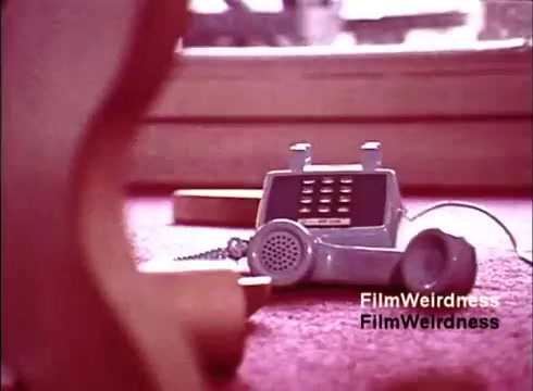 The still features a 1970s-style telephone, prominently displayed on a carpeted surface, with a close-up perspective. The phone has a classic design, showcasing a keypad and a receiver, capturing the nostalgia of the Bell System era. The soft color palette and lighting contribute to a vintage feel, emphasizing a sense of history and innovation in telecommunications. The surrounding area suggests a casual setting, adding to the personal connection to the technology of that time.