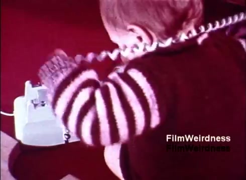 The still from the 16mm film 'Bell Performance' features a young child interacting with a classic telephone. The child is dressed in a striped sweater and is engaged with the phone cord, which adds a nostalgic touch to the scene. The background hints at a cozy setting, indicative of the domestic use of telephones in that era. Overall, the image evokes a sense of nostalgia related to the Bell System and its impact on communication during the 1970s.