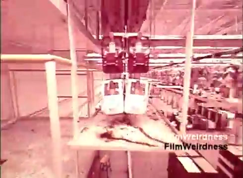 The still captures a moment from the film 'Bell Performance,' showcasing the manufacturing process of telephones in a factory setting. It features assembly line equipment, with two devices suspended above a conveyor. The scene reflects the era's industrial approach to telecommunications, accompanied by a nostalgic aesthetic typical of 1970s films. The background hints at various parts and materials, emphasizing the scale and complexity of telephone production during that time.