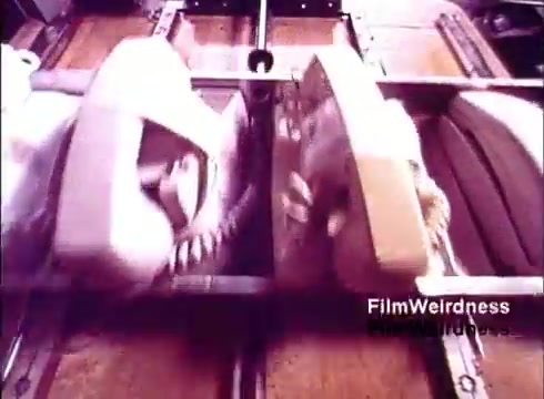 The still captures a close-up view of two vintage telephones on a production line, likely from the 1970s, showcasing the manufacturing process. The phones are cream and beige, highlighting the design of that era. The setting appears industrial, with machinery and assembly components in the frame, emphasizing the nostalgia associated with Bell System's telephone creation. The overall tone is reminiscent of the technological advancements of the time.