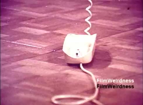 The 16mm still from 'Bell Performance' features a close-up of a white telephone on the floor, with a coiled cord extending from it. The background appears to have a wooden or textured surface. The image likely captures the essence of telecommunications nostalgia from the 1970s, highlighting the simplicity and design of that era's technology. The overall aesthetic suggests a focus on the manufacturing or assembly process of telephones.