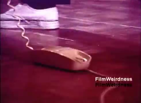 The still from 'Bell System Film from the 1970s' features a close-up of a telephone from that era. It shows a beige phone on a wooden floor, with a curly cord connecting it to a base. In the background, a person's foot in a sneaker is visible, suggesting a casual setting. This image captures a moment in the manufacturing or showcasing of telephones, evoking nostalgia for the Bell System's influence on communication technology during that time.