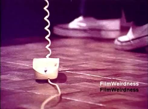 The still from the film 'Bell Performance' showcases a retro telephone on a wooden floor, featuring a coiled cord. In the background, feet in casual footwear are slightly visible, suggesting a domestic setting. The image evokes nostalgia for the era of the Bell System, highlighting the simplicity and design of telecommunication devices from the 1970s.