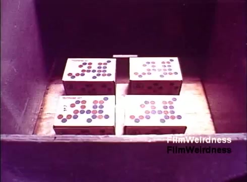 The still from 'Bell Performance' features a close-up of several boxes placed inside a wooden container. Each box is adorned with colorful dots, possibly indicating various components or features related to telephone manufacturing. The overall aesthetic reflects a nostalgic 1970s vibe, capturing a moment in the historic production of telecommunication devices. The lack of titles suggests an observational style, focusing on the process rather than a narrative.