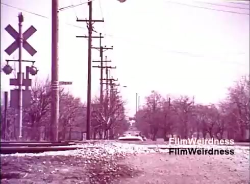 The still from the 'Bell System Film from the 1970s' shows a tranquil scene along a rural road, featuring a set of railway tracks. Telephone poles line the street, suggesting the presence of telecommunication infrastructure. The landscape appears partially barren, likely reflecting the time of year, with sparse trees on either side. The overall muted colors evoke a sense of nostalgia, emphasizing the simplicity and functionality of mid-20th-century American life. The film seems to capture the essence of Bell System's operations and community impact during that era.