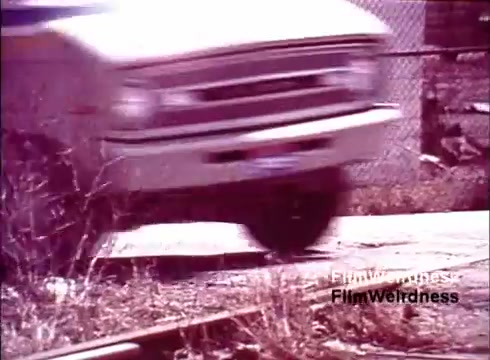 The still from 'Bell Performance' showcases a vintage scene featuring a vehicle, likely a truck, speeding past a set of railway tracks. The focus is on the motion of the truck, with a blurred effect emphasizing speed. The background includes an industrial setting, hinting at a telephone manufacturing environment. The overall color tone is reminiscent of film from the 1970s, evoking nostalgia for that era in telecommunication history.