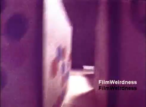 The still from the 'Bell System Film from the 1970s' captures an intriguing glimpse into the manufacturing process of telephones. The scene features a corridor or pathway lined with large, possibly colorful boxes or equipment, set in a warm hue, indicative of the film's age and nostalgic aesthetic. The composition suggests movement, leading viewers to feel as though they’re navigating through the space where telephones were once produced. Overall, it evokes a sense of curiosity about the technology and era.