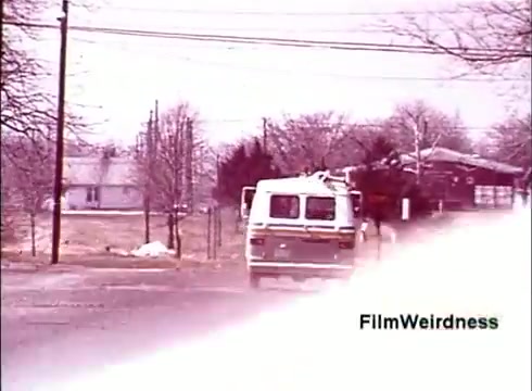 The still from the 16mm film 'Bell System Film from the 1970s' depicts a scene with a van driving down a road lined with bare trees and houses in the background. The color palette seems muted, evoking a nostalgic feel. The van likely represents Bell System's operational presence, while the residential setting hints at its connection to everyday life and telecommunications development during that era. The image focuses on the simplicity and functionality associated with Bell's services.
