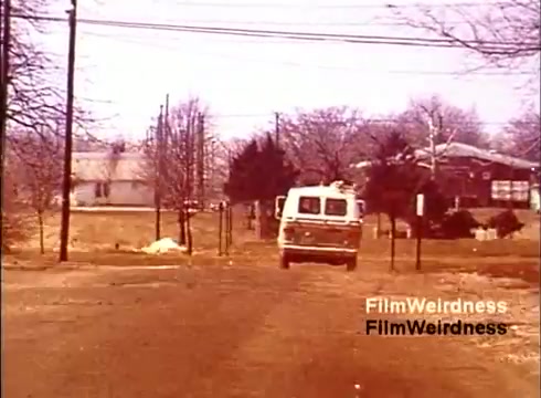 The still showcases a scene from the 1970s film 'Bell Performance,' featuring a white van driving down a rural road lined with bare trees. The surrounding landscape suggests a quiet, suburban environment, with houses visible in the background. The overall aesthetic captures a nostalgic era, likely highlighting the telecommunications industry's evolution, including the manufacturing and distribution of telephones. The scene evokes a sense of simplicity and the foundational aspects of communication technology during that time.