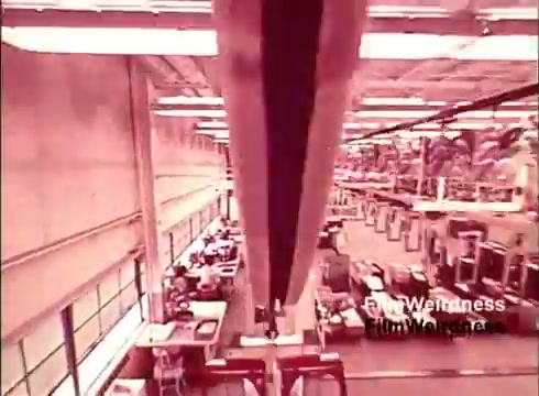 The still from the 16mm film 'Bell Performance' showcases a factory setting, likely within a Bell System manufacturing facility. It captures an overhead view of production lines where telephones are being assembled. The environment features organized rows of workers and machinery, highlighting the industrial processes of the 1970s. The color tone suggests a vintage feel, adding to the nostalgia associated with the Bell System. The absence of titles reinforces its documentary style, focusing on the manufacturing process itself.