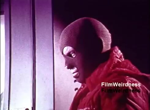 The still from the 16mm film 'Bell Performance' features a figure wearing a ski mask and a red jacket, standing near a door. The context suggests a mix of nostalgia for Bell System, possibly highlighting the telecommunications industry while incorporating a mysterious or dramatic element. The film appears to explore the creation and assembly of telephones, providing a unique glimpse into the era’s technological landscape.