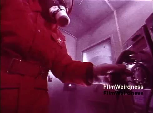 The still from 'Bell Performance' captures a worker in a bright red suit, complete with a gas mask, interacting with a rotary telephone in a lab-like setting. The overall color tone is tinted purple, which adds a surreal quality to the image. This frame likely highlights the production and testing process of telephones in the Bell System, evoking a sense of nostalgia for the era's technology and safety protocols.