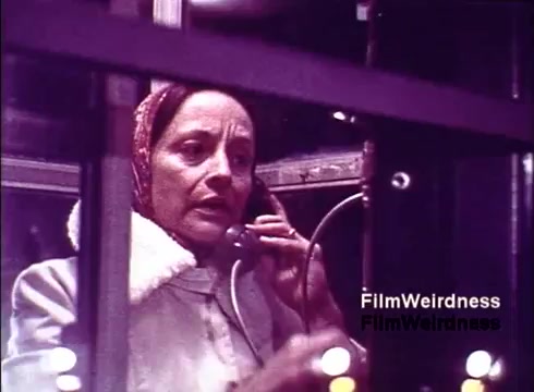 The still from 'Bell System Film from the 1970s' features a woman inside a telephone booth, engaged in a phone call. She is wearing a headscarf and a coat, conveying a sense of nostalgia typical of that era. The image captures a moment of communication technology from the past, highlighting the everyday use of telephones. The overall atmosphere reflects a blend of personal interaction and the technological landscape of the time.