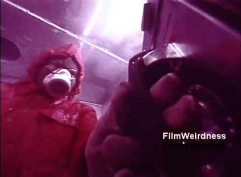 The still from the 1970s film 'Bell Performance' shows a worker in a bright red jacket, possibly in a manufacturing or production environment. The worker appears to be holding a cup, likely taking a break or observing the process. The scene suggests a focus on telephone manufacturing, highlighting the behind-the-scenes work in the Bell System. The lighting has a pinkish hue, adding a nostalgic feel to the industrial setting.