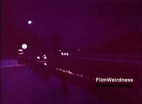 The still captures a nighttime scene, possibly depicting a road or highway setting with vehicles illuminated by headlights. The overall color tone leans towards a purple hue, giving it a nostalgic, atmospheric feel. A sign can be seen in the foreground, and the film seems to exude a vintage quality that evokes memories of the 1970s. This visual aligns with the theme of the Bell System, hinting at a focus on technology and transportation during that era.