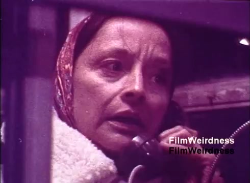 The still from the 'Bell System Film from the 1970s' features a woman with a serious expression, holding a telephone while in a seemingly working environment. She is dressed modestly, with a scarf covering her hair. The film appears to focus on the context of telephone manufacturing or usage, capturing a moment filled with nostalgia related to the Bell System's operations. The overall tone suggests a documentary style, emphasizing the human element in telecommunications.