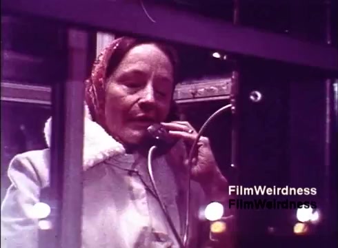 The still features a woman inside a telephone booth, engaged in a conversation on a traditional telephone. She appears to be wearing a light-colored coat with a fur collar and a scarf, suggesting a vintage style typical of the era. The background shows the booth's interior, illuminated by soft lighting, emphasizing a sense of personal connection and nostalgia associated with telephone technology from the 1970s. This moment captures the essence of communication during that time period, reflecting the significance of the telephone in everyday life.