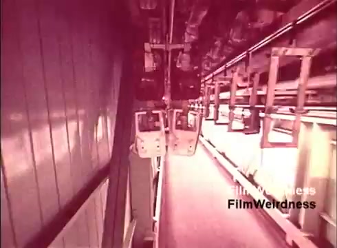 The 16mm still from the 'Bell System Film from the 1970s' depicts a factory setting where telephones are being manufactured. The image gives a glimpse into the assembly process, with various components moving along a production line. The lighting and color give it a nostalgic feel, indicative of the era. It captures a moment of technological history, reflecting the significance of telecommunications in daily life during that time.