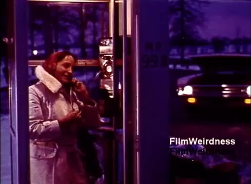 The still from the 1970s film 'Bell Performance' showcases a woman using a payphone, dressed warmly in a light-colored coat and fur collar. The scene is illuminated in a purple hue, adding a nostalgic feel. In the background, a car is visible, suggesting a winter setting with bare trees. This image captures the everyday use of telephone technology during that era, encapsulating a moment of communication within the context of Bell System's impact on society.