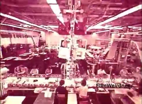The 16mm still from 'Bell System Film from the 1970s,' titled 'Bell Performance,' showcases a bustling factory environment where telephones are manufactured. The scene captures workers engaged in assembly tasks, with various parts and tools spread across a conveyor belt. The warm color tone enhances the nostalgic feel, highlighting the manual craftsmanship involved in creating telecommunication devices during that era. The overall atmosphere reflects the industrial efficiency of the Bell System at the time.