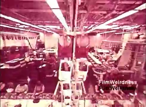 The still from the 'Bell System Film from the 1970s' shows an industrial scene likely within a telephone manufacturing facility. The image captures a bustling workspace filled with machinery and assembly lines, emphasizing the production aspect of telephones. Workers can be seen engaged in various tasks, showcasing the manufacturing process. The overall atmosphere reflects a sense of nostalgia for the era, particularly in relation to the Bell System's legacy in telecommunications. The color tone appears warm and dated, enhancing the vintage feel of the film.