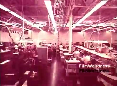 The still from 'Bell Performance' depicts a busy manufacturing facility where telephones are being assembled. The image features numerous workers engaged in various tasks along long assembly lines, surrounded by machinery and equipment. The lighting has a pinkish hue, giving the scene a nostalgic quality that reflects the era of the 1970s. The overall atmosphere suggests a blend of craftsmanship and industrial efficiency characteristic of Bell System operations during that time.
