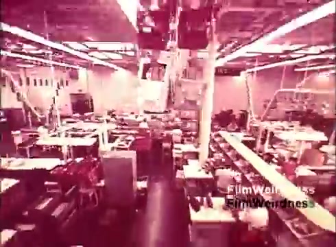 The still from 'Bell System Film from the 1970s' captures an expansive manufacturing facility where telephones are being produced. The image showcases numerous workstations filled with equipment and tools, reflecting the intricate processes of telephone assembly. The lighting is bright, creating a lively and industrious atmosphere, typical of factory environments from that era. The overall aesthetic exudes a sense of nostalgia for Bell System technology and innovation.