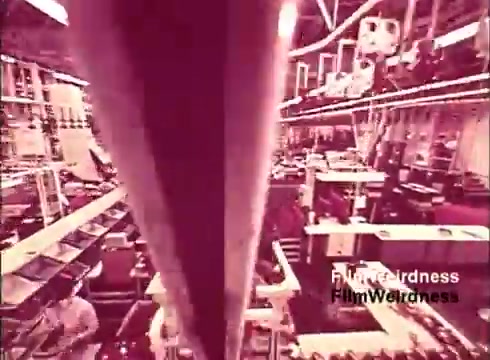 The still from the 16mm film 'Bell Performance' shows an industrial setting, likely a telecommunications manufacturing facility. The image captures a busy assembly line with workers engaged in the production of telephones. The environment appears organized with equipment and materials arranged methodically, highlighting the process of telephone creation. The film evokes a sense of nostalgia for the Bell System, reflecting on the technology and labor involved in telecommunications during the 1970s.